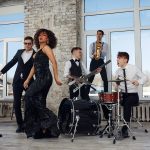 Jazz band performing with a singer and musicians playing saxophone, drums, and guitar in a stylish loft with large windows.
